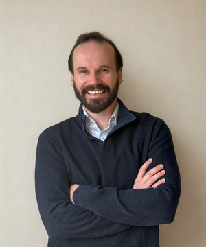 Man with a beard wearing a dark sweater on a beige background.