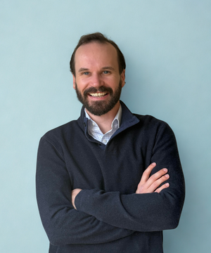 Man with a beard wearing a dark sweater on a blue background.